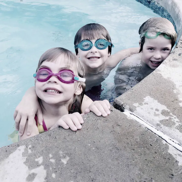 Three smiling children in swim goggles are enjoying a swim in a pool, with one girl hugging another by the edge.