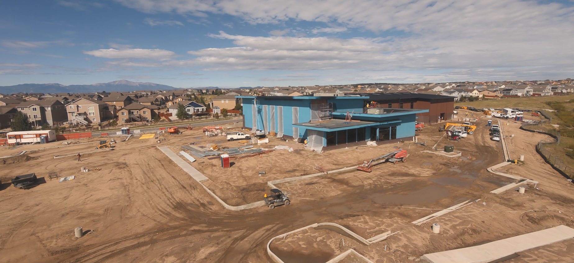 Construction site with a blue building, vehicles, and equipment, surrounded by residential houses under a partly cloudy sky.