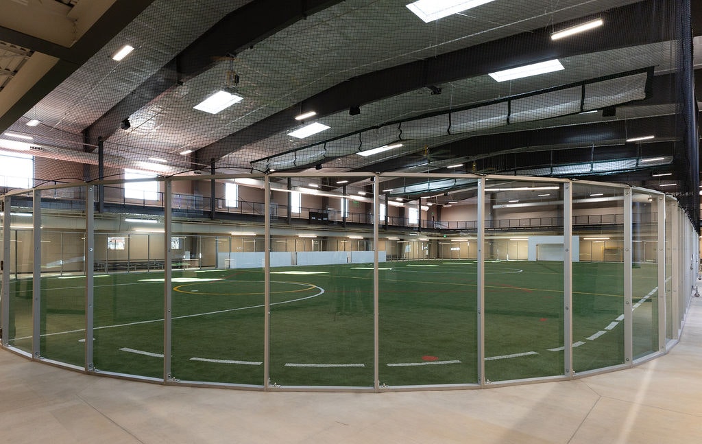 The image shows an indoor sports facility with a glass-enclosed turf field, designed for soccer or similar activities.