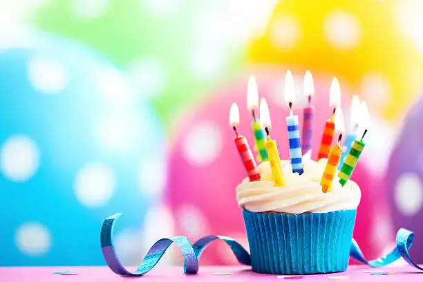 A colorful cupcake topped with frosting and birthday candles, set against a vibrant, polka-dotted background.