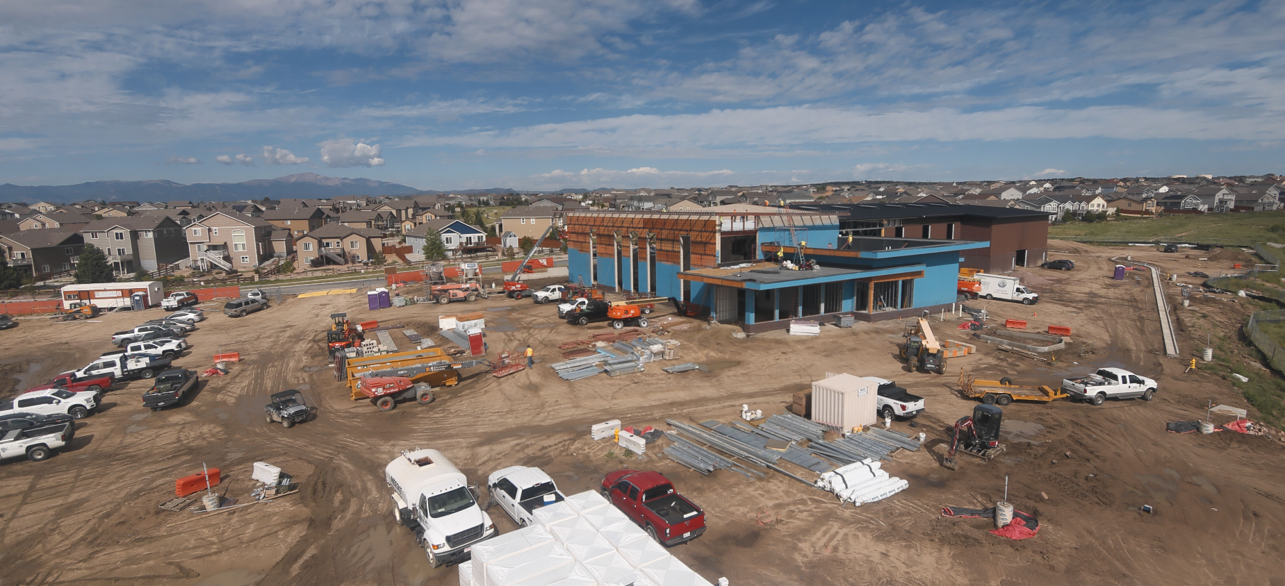 Construction site with vehicles, equipment, and building materials. Houses in the background under a blue sky.
