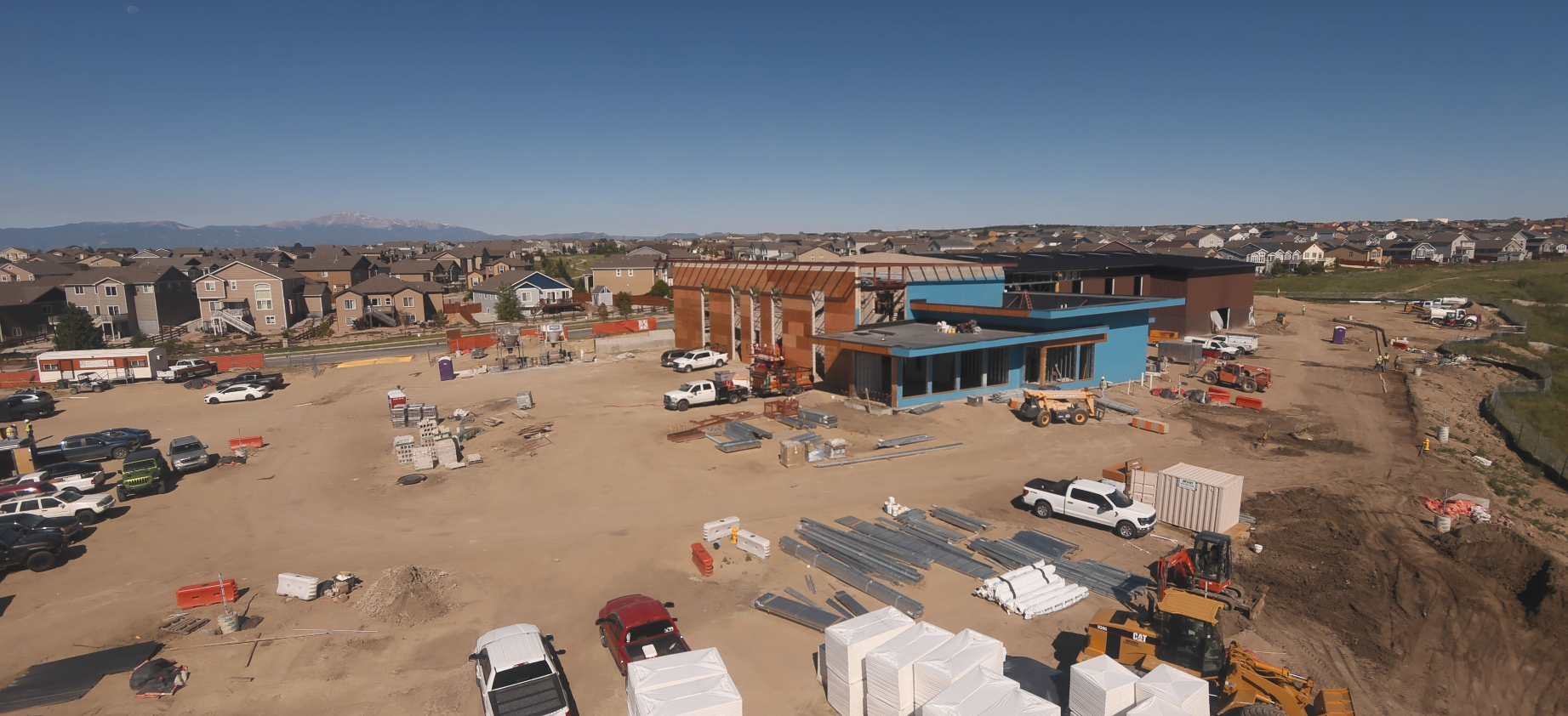 Construction site with vehicles, building materials, and partially completed structures; residential neighborhood in the background.