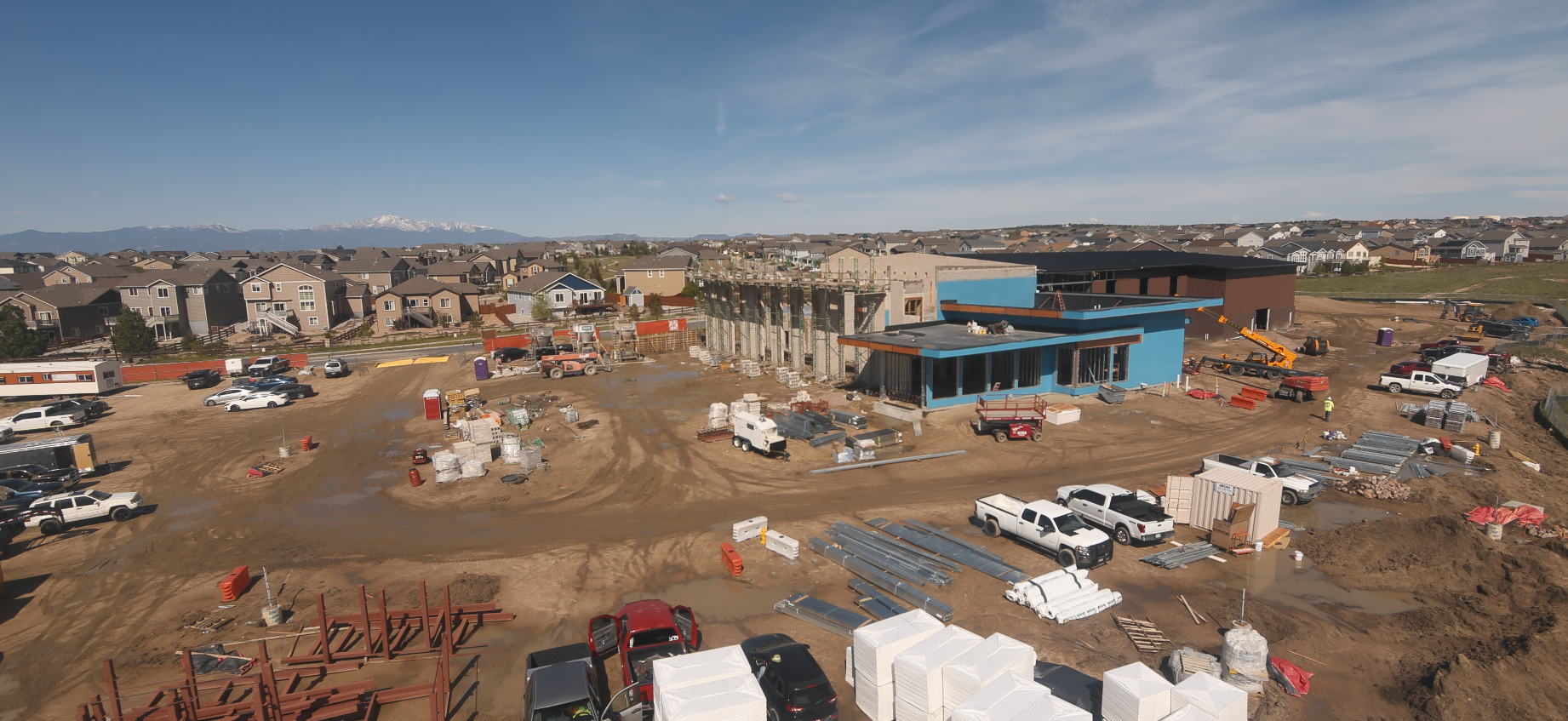 A construction site with building materials, vehicles, and partially built structures, surrounded by residential houses and mountains in the distance.