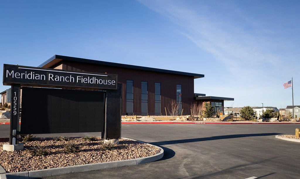 The image shows the Meridian Ranch Fieldhouse, a modern building with large windows, landscaping, and an American flag nearby.