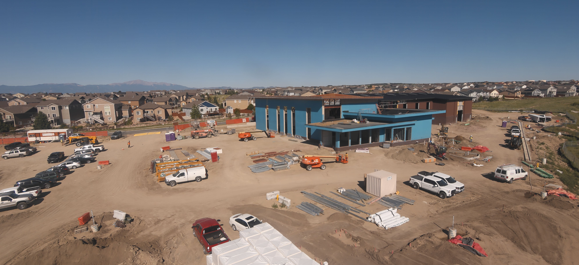 Construction site with vehicles, building materials, and a blue structure. Residential area and mountains in the background.
