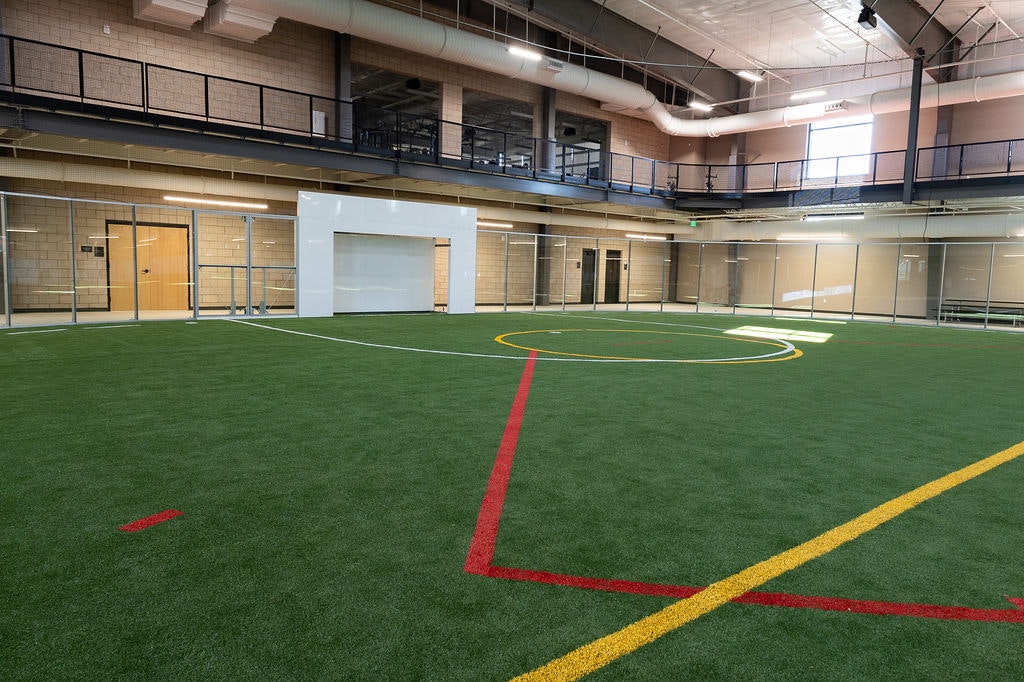The image shows an indoor sports field with artificial turf, marked lines, and a goal area, surrounded by glass walls.