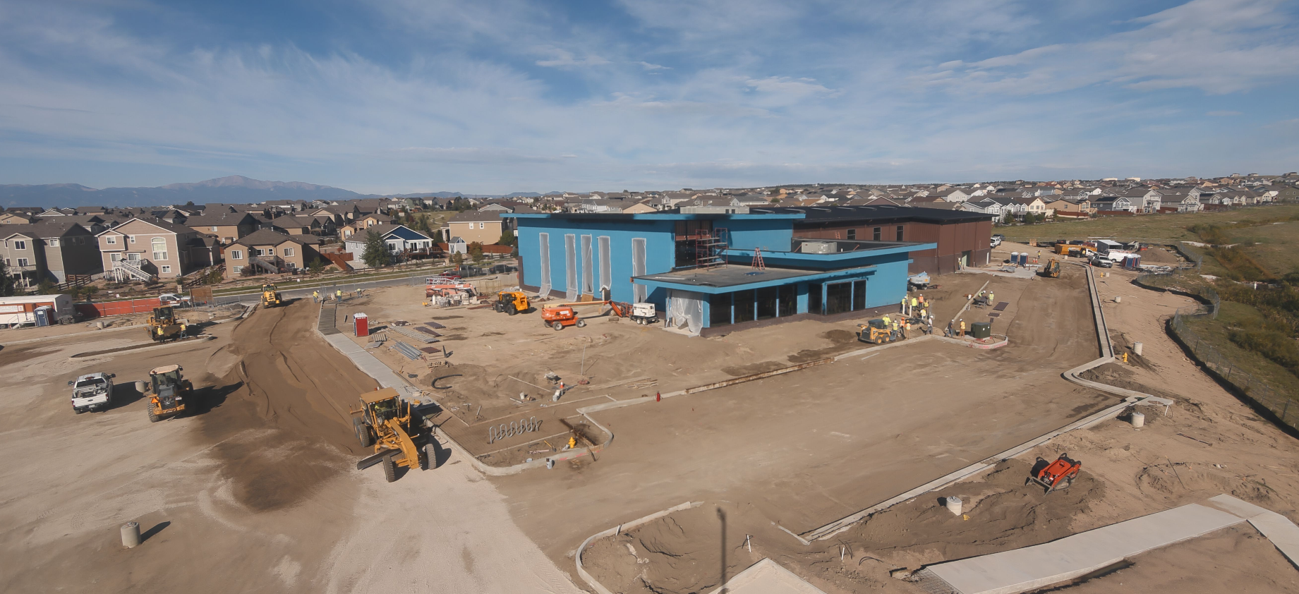Construction site with heavy machinery, workers, and a blue building in a suburban area. House neighborhood in the background.