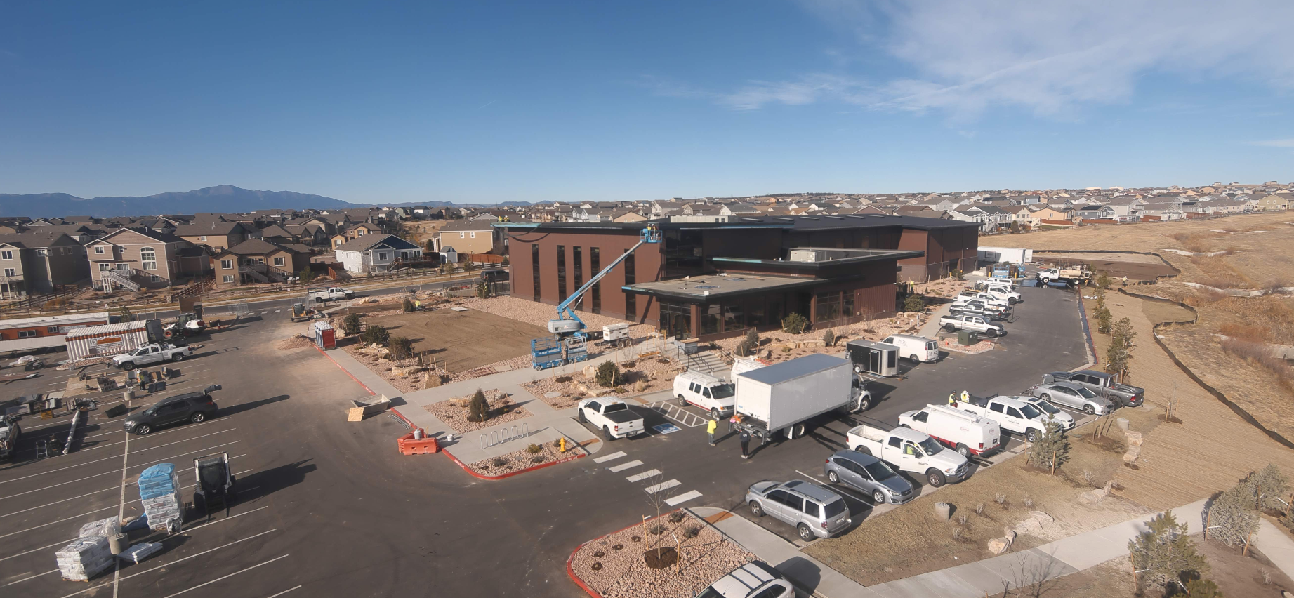 Construction site with a large building, vehicles, and workers; residential area and mountains in the background.