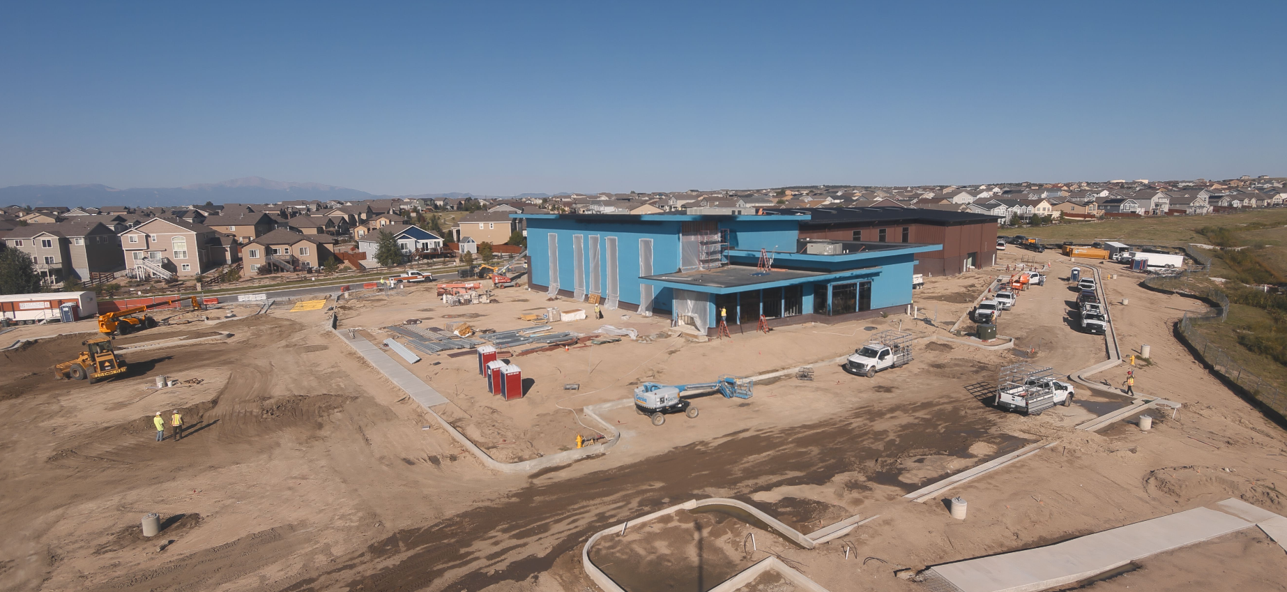 Construction site with a blue building, trucks, equipment, and workers. Nearby residential area under clear sky.