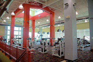 The image shows a modern gym interior with various exercise machines and colorful decor, featuring large windows and bright lighting.