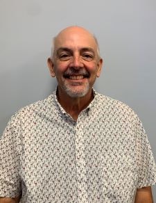 A smiling person wearing a patterned shirt stands against a plain background.