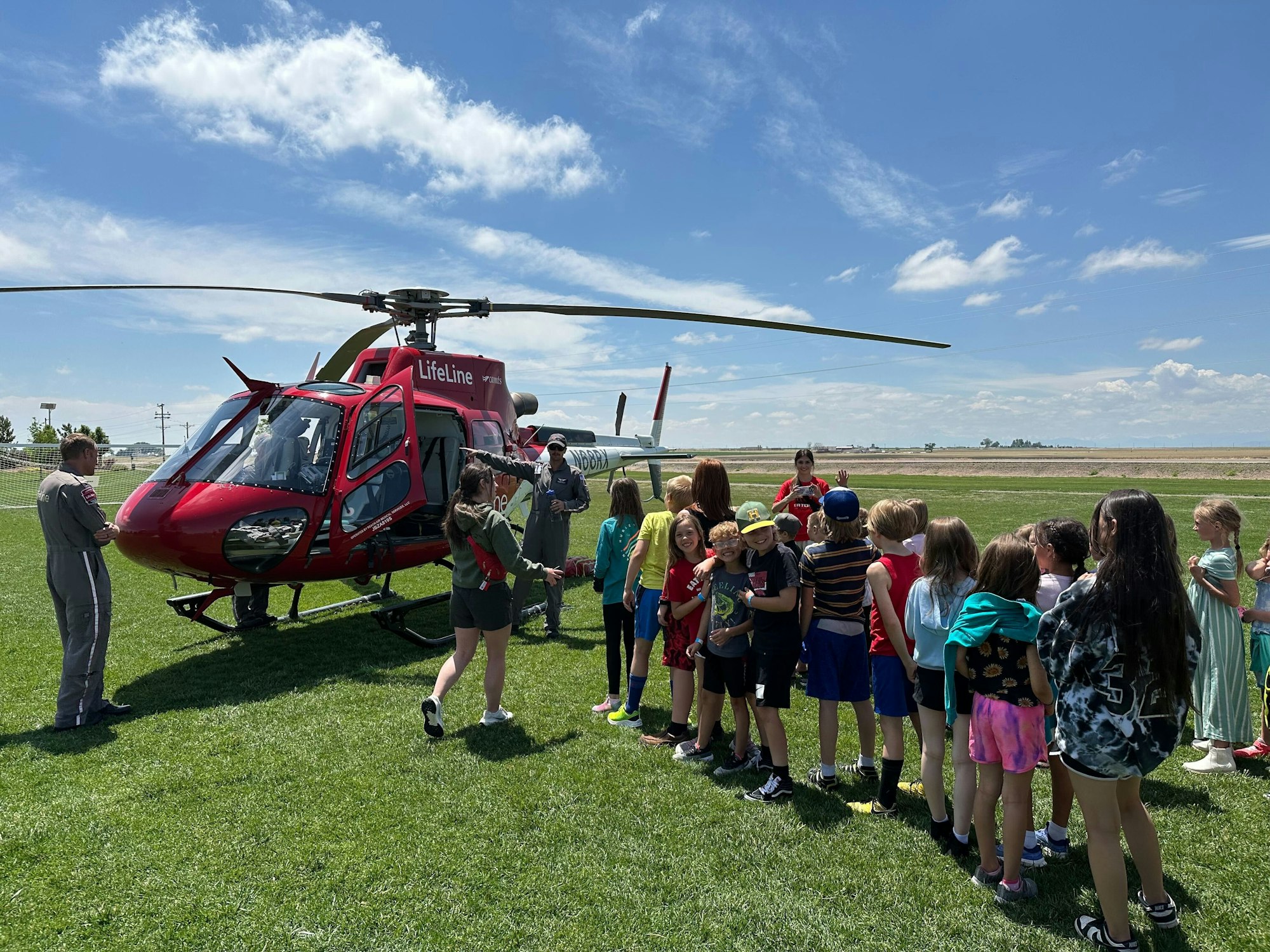 A group of children stands near a red helicopter on grass, with adults supervising.