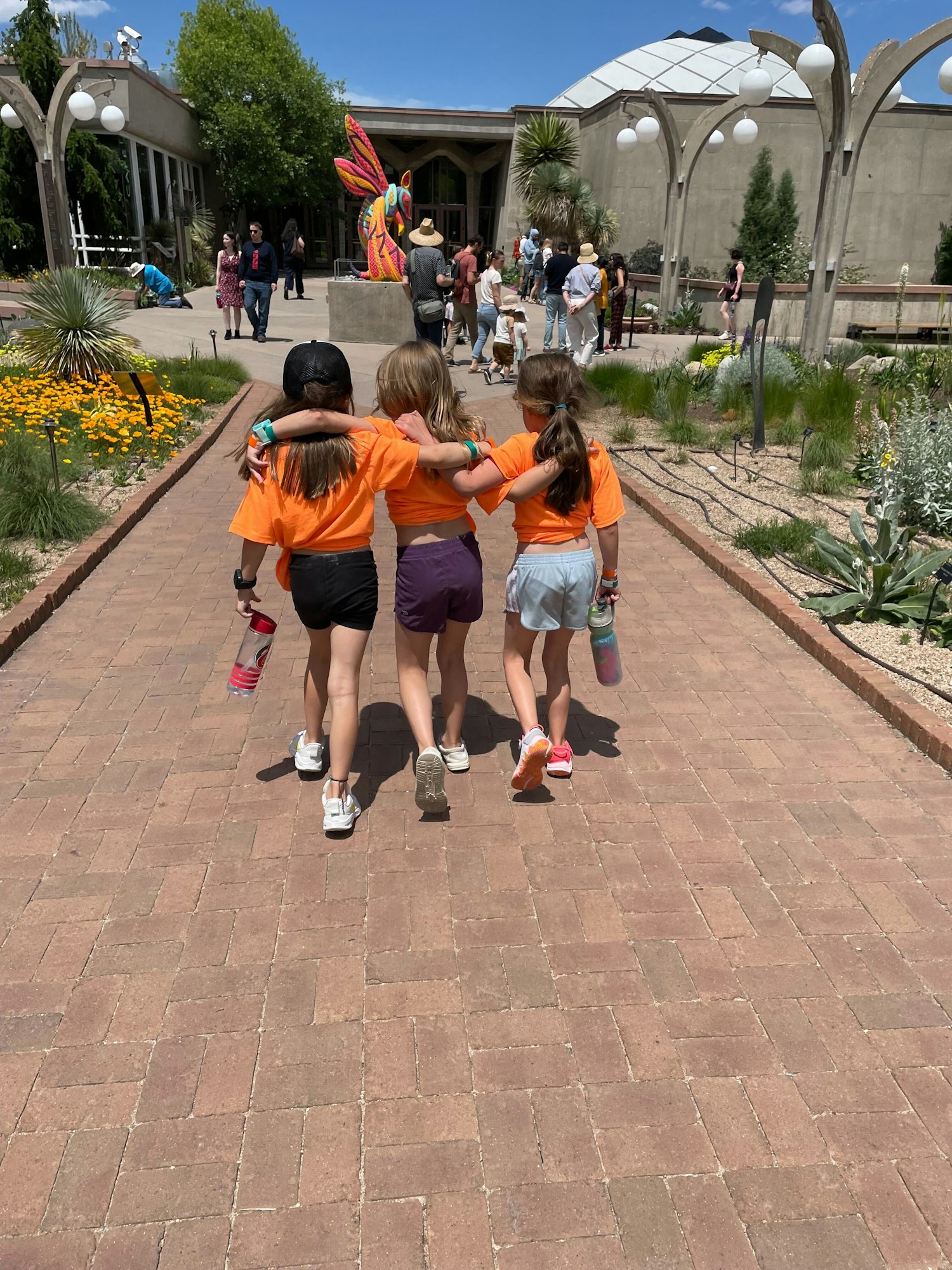 Four children in orange shirts walk arm in arm down a path, with people and greenery around them.