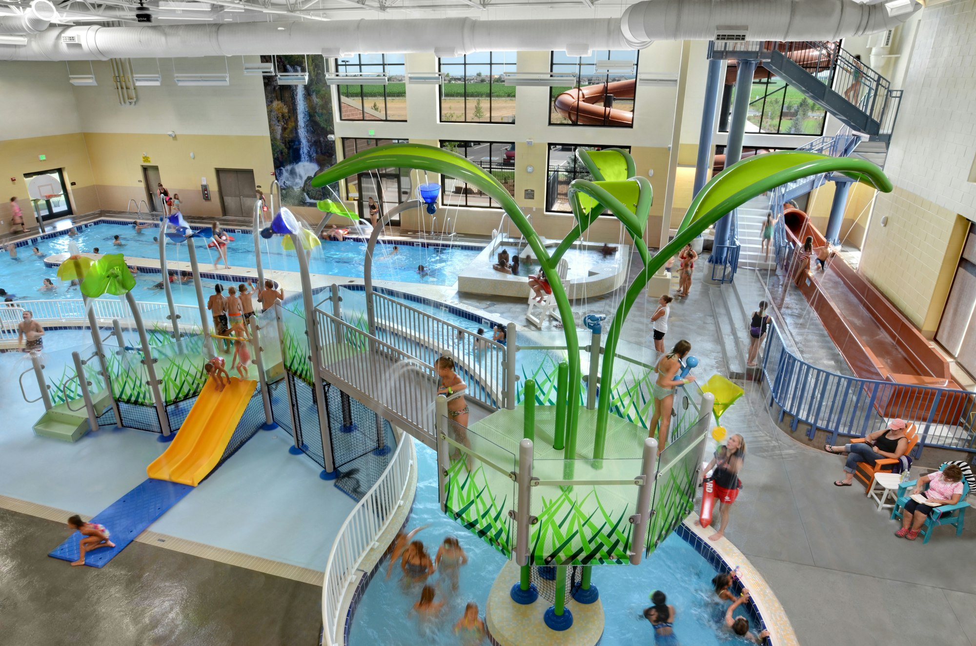 Indoor pool with people swimming, slides, play structures, and spectators relaxing.