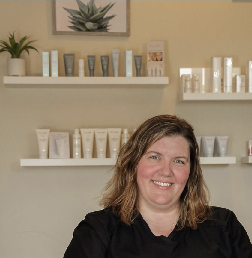 A woman smiling in a skincare clinic with product shelves and a potted plant on the wall.