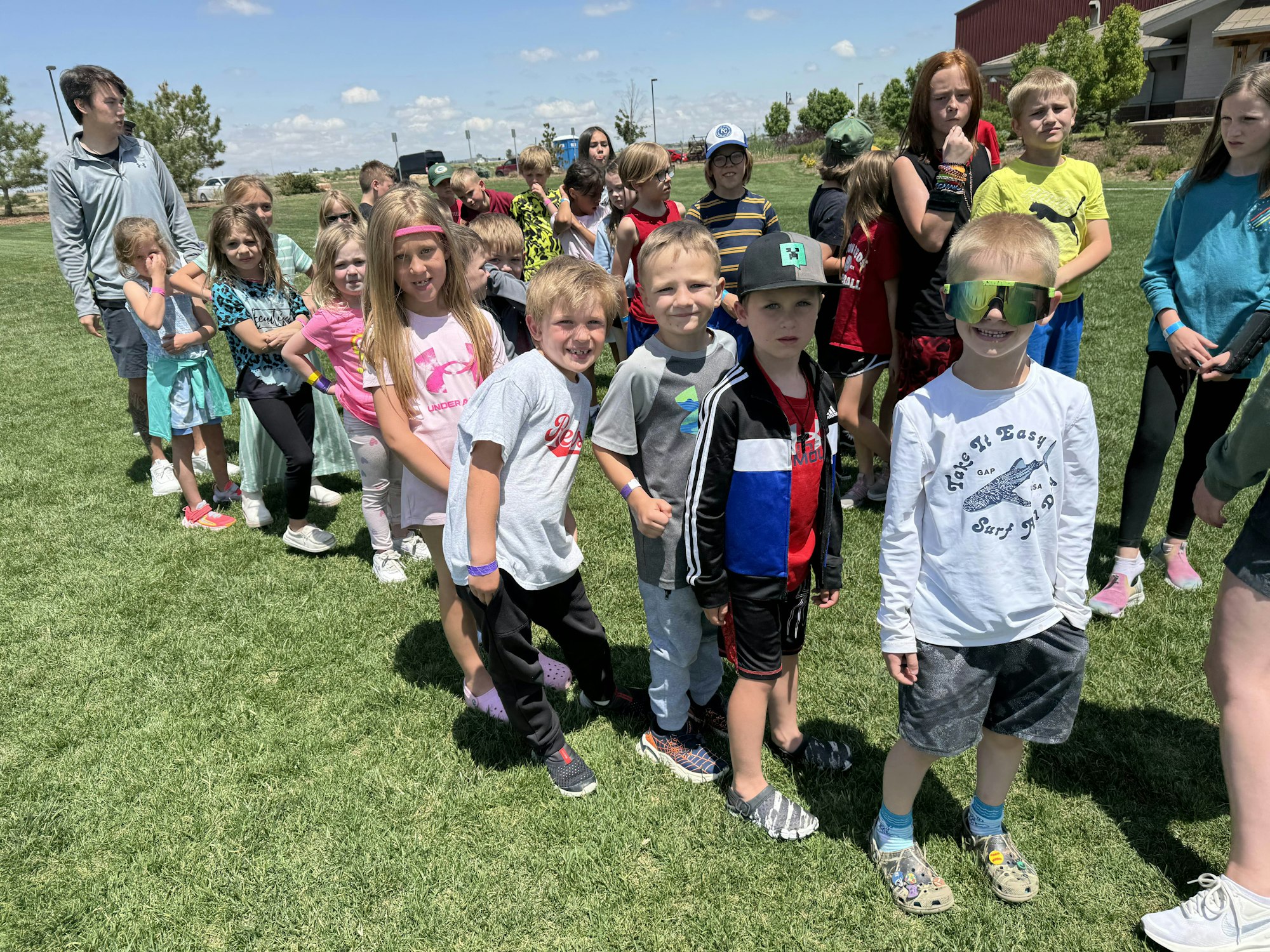 A group of children and a few adults stand in a line outdoors on a grassy field under a sunny sky.