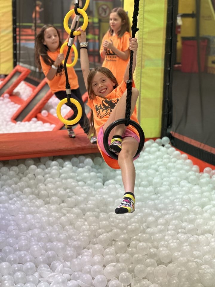 Kids in orange shirts playing on swinging rings above a ball pit.
