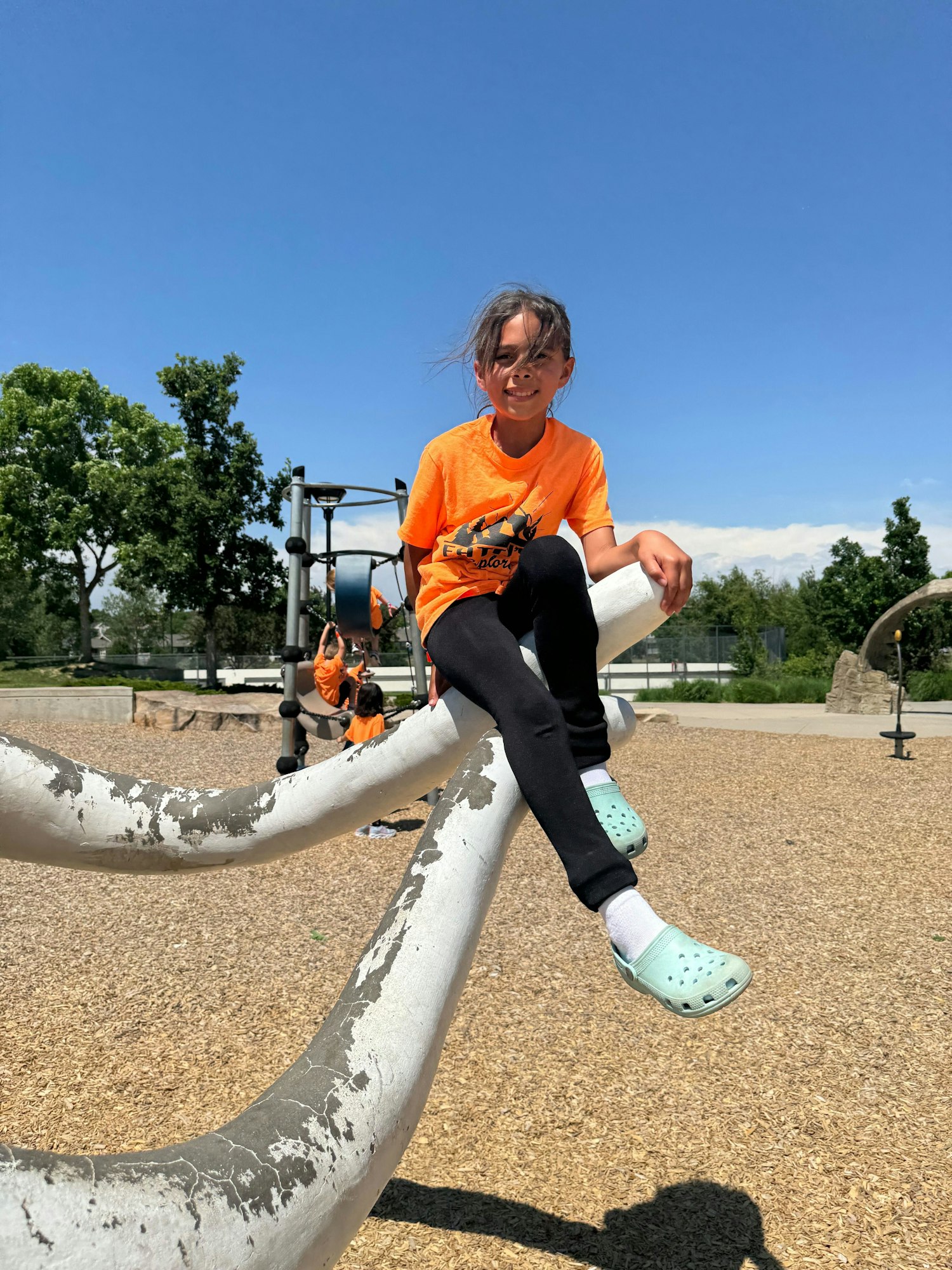 Child in an orange shirt sitting on a playground structure, sunny day, blue sky.