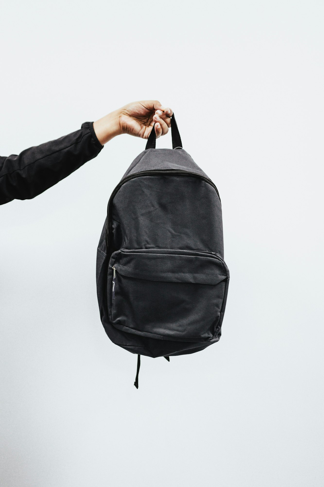 A person holds a black backpack by its strap against a light background.