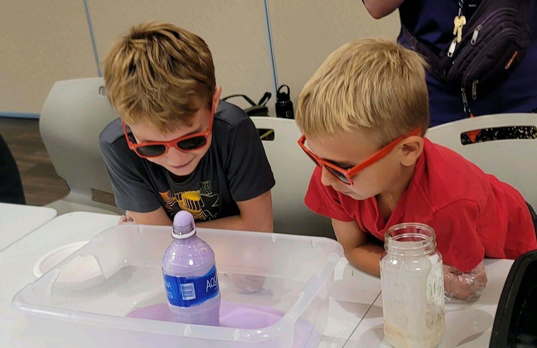 Two kids with red sunglasses watch a bottle with purple liquid in a tray.
