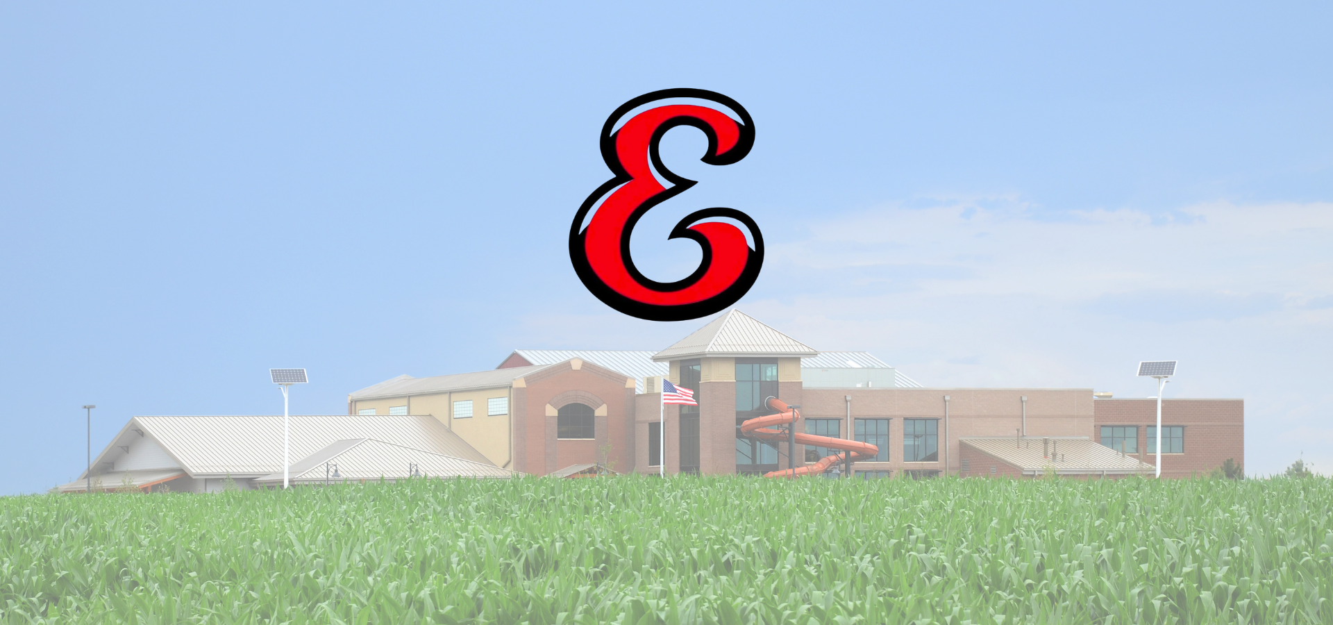The image features a large building in a field, topped with a prominent ampersand symbol against a blue sky.