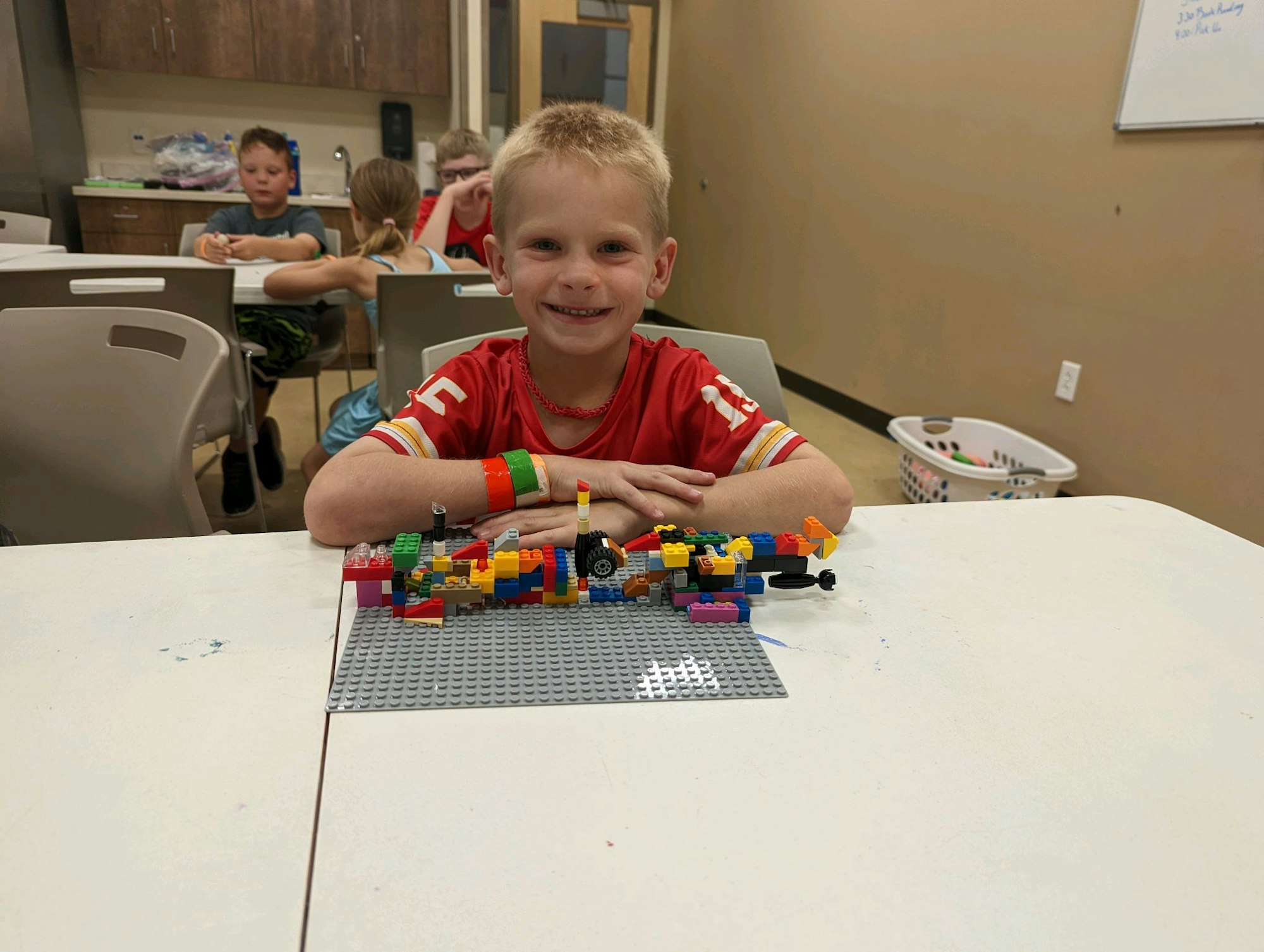 A child in a red shirt smiles, sitting at a table with a LEGO creation. Other kids are visible in the background.