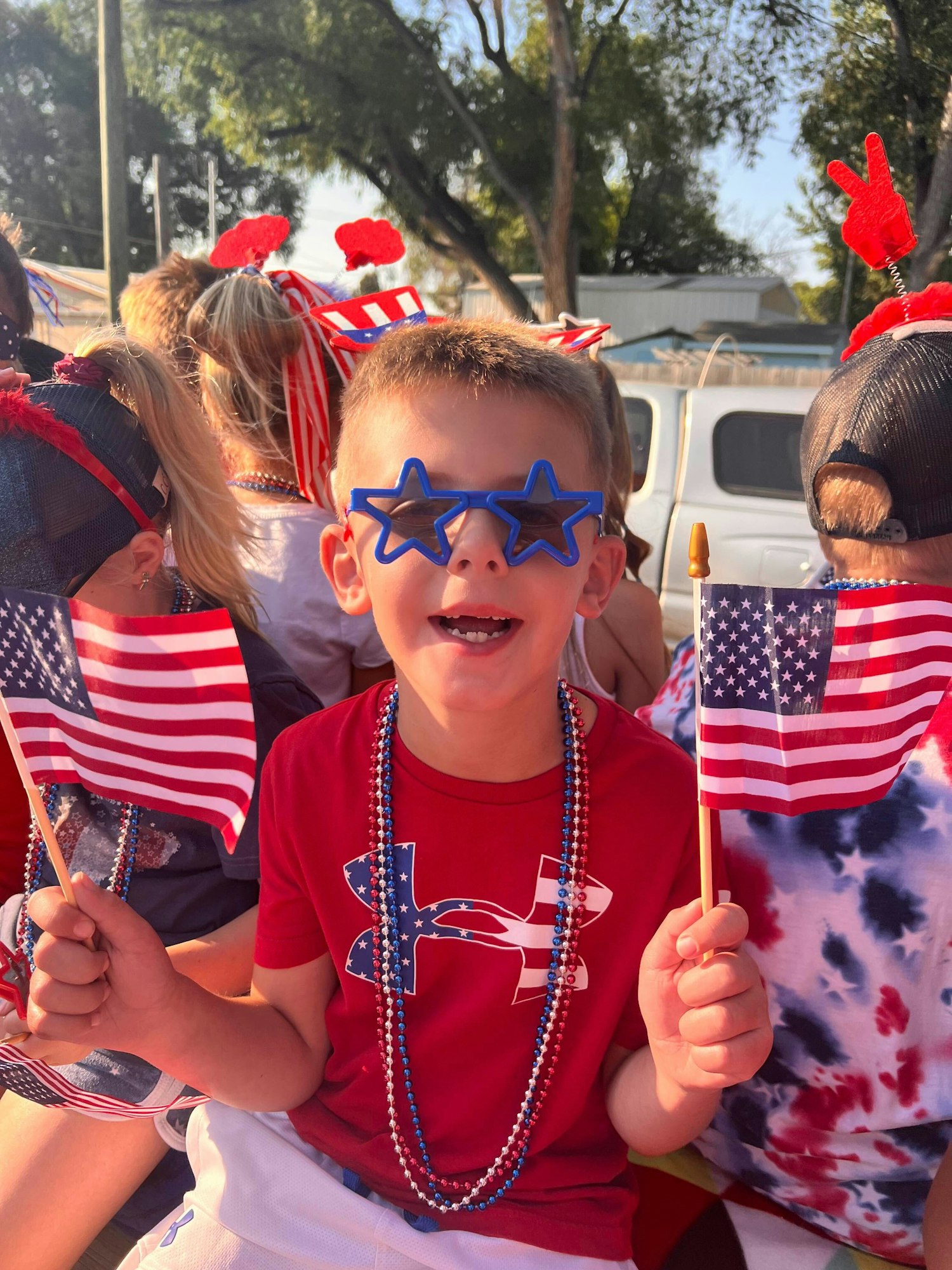 A child wearing star-shaped sunglasses, holding American flags, and dressed in red, white, and blue during a festive gathering.