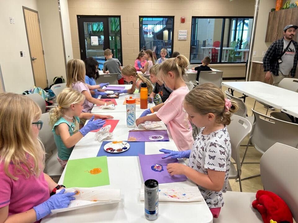 Children in a room doing crafts at tables with paper and paint, wearing gloves.