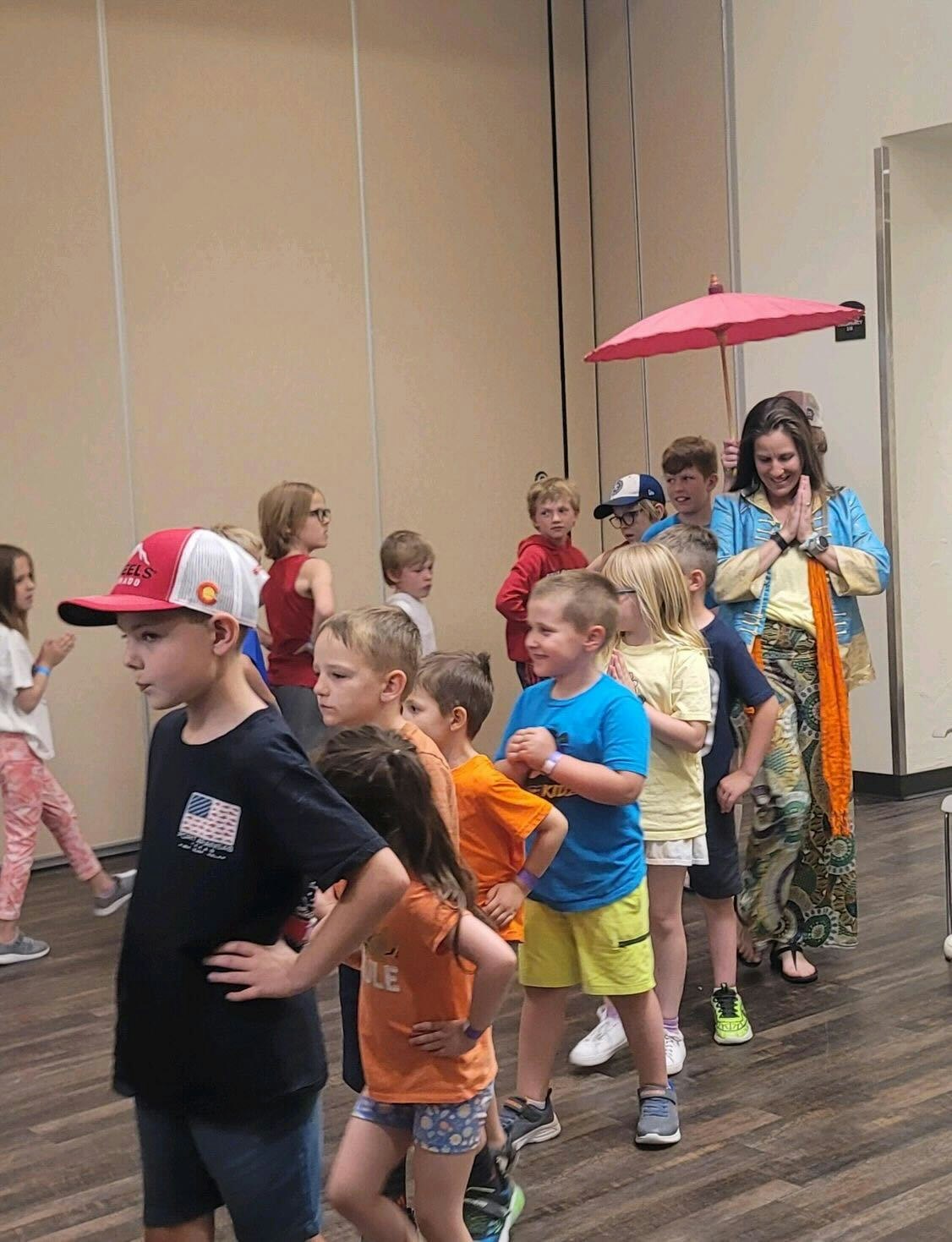 Children are standing in line indoors, with a person holding a pink umbrella in the background.