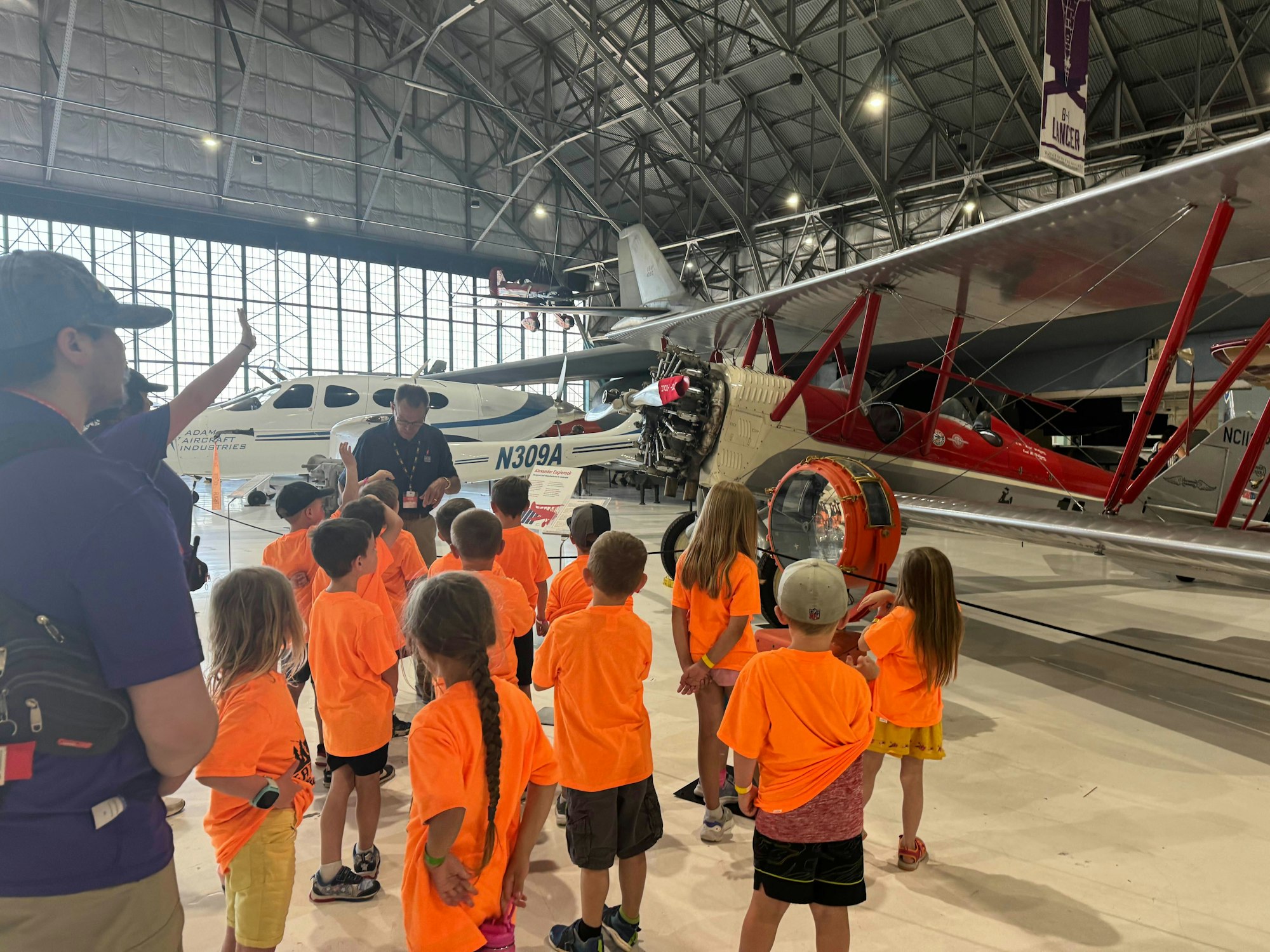 Group of kids in bright orange shirts on a tour at an airplane museum, looking at aircraft.