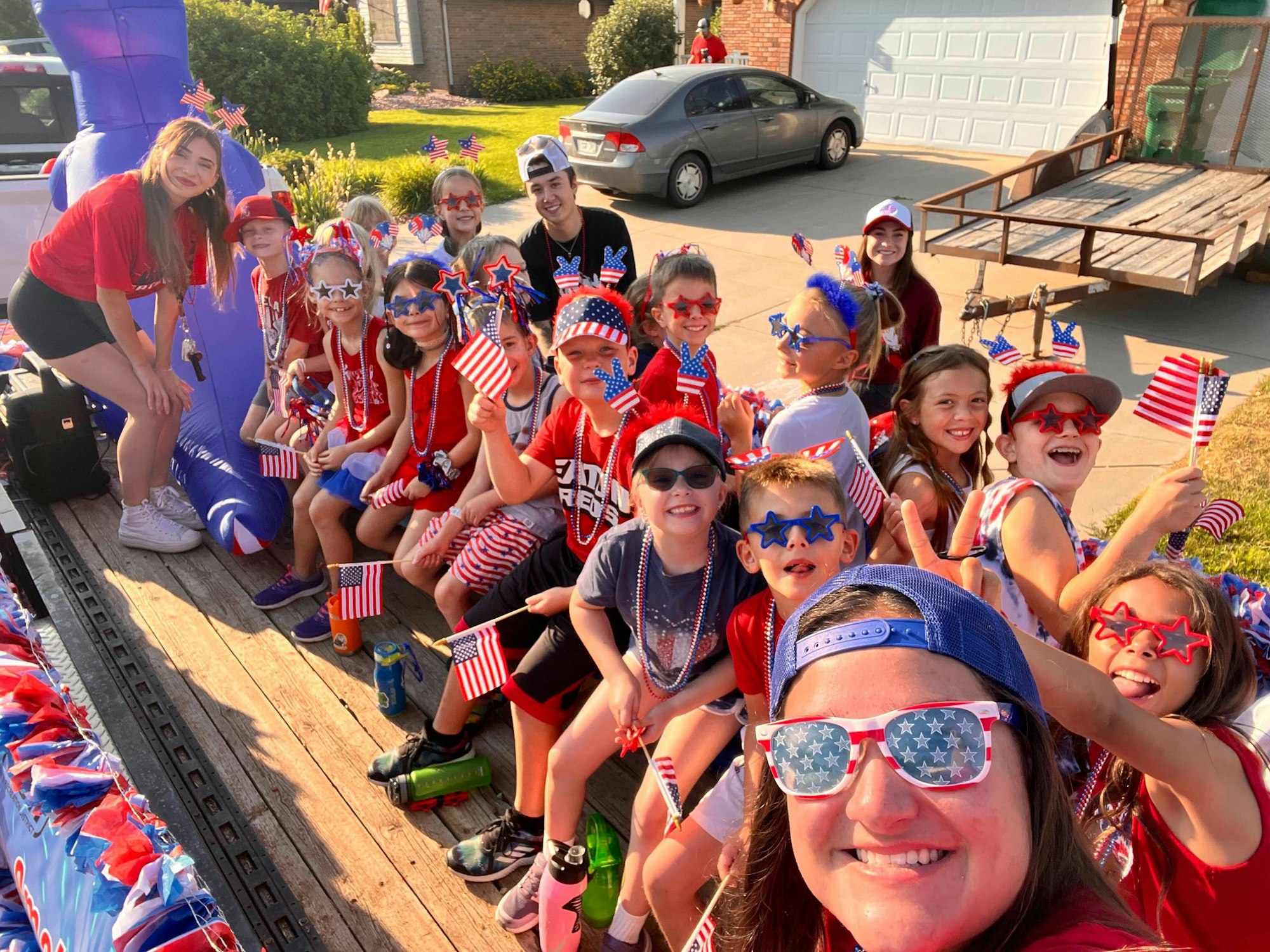 A group of people, mostly kids, wearing red, white, and blue and holding American flags, celebrate outdoors.