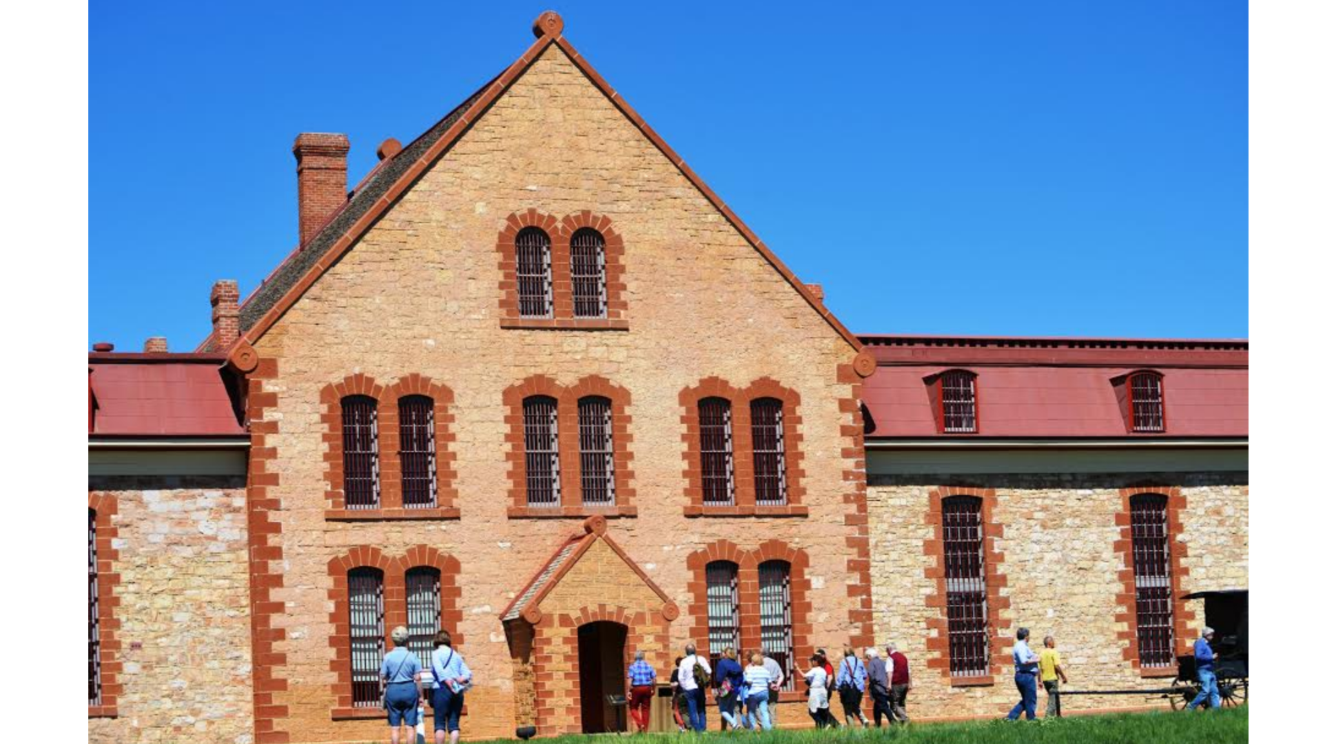 A historic building with large windows, likely an old prison, and a group of visitors exploring the site under a clear blue sky.