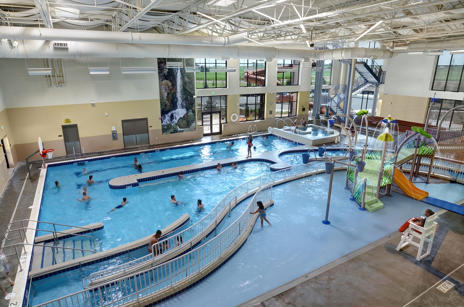 The image shows an indoor swimming pool complex with people swimming, water slides, and play equipment for kids.