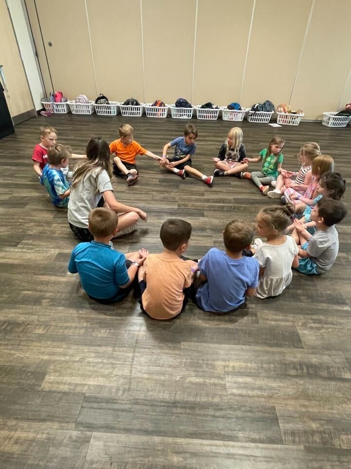 Children sitting in a circle on the floor, holding hands, in a room with baskets along the wall.