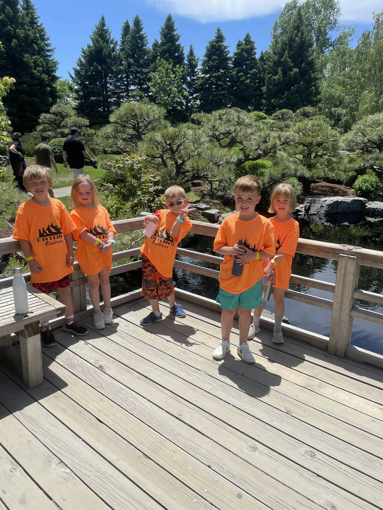 Five kids in orange shirts stand on a wooden deck by a pond, surrounded by trees.