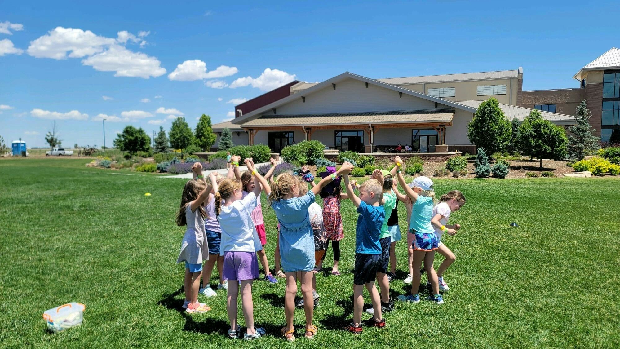 Children holding hands in a circle outdoors, grassy area, clear sky, building in the background.