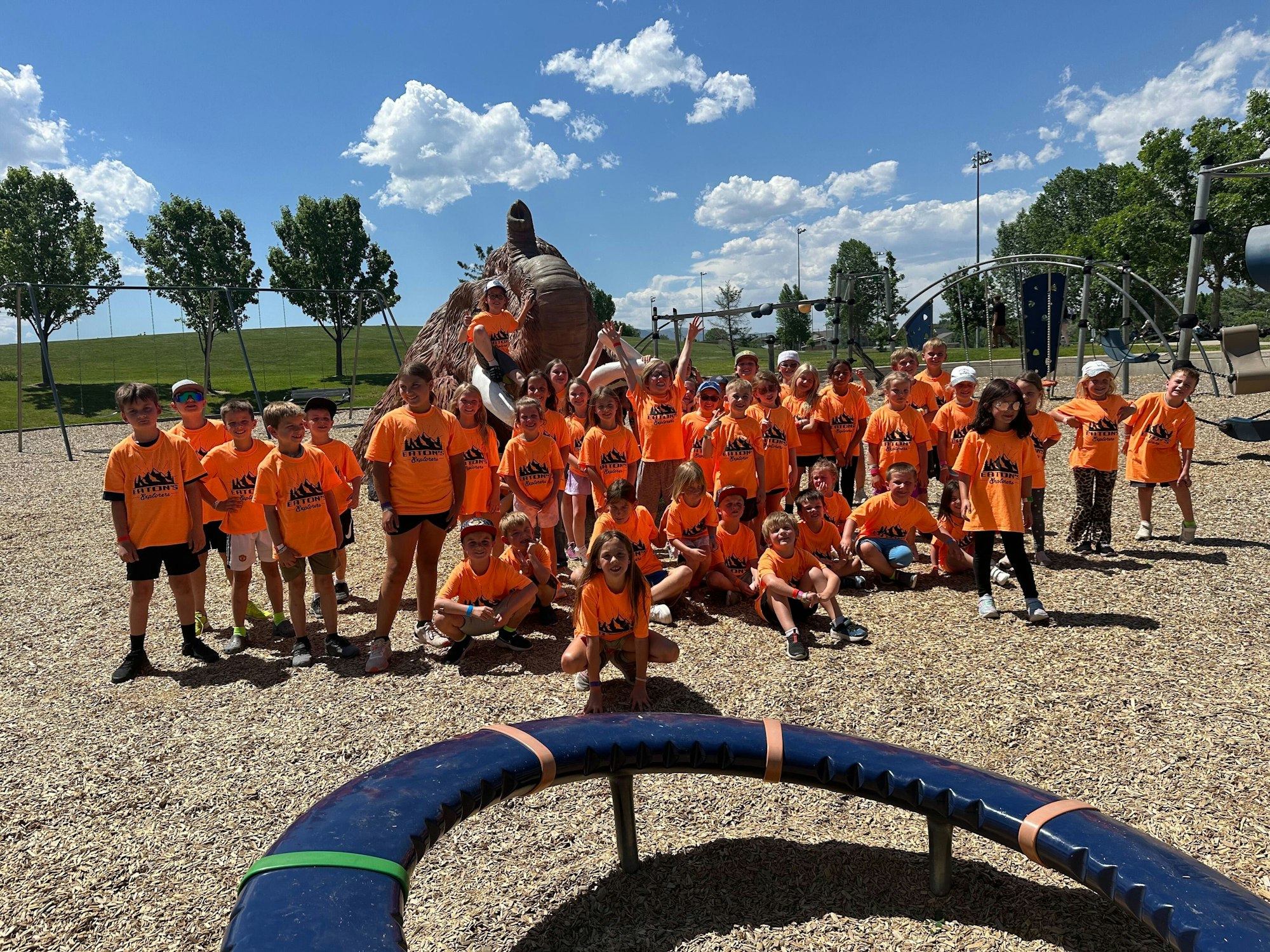 A group of kids in orange shirts posing at a playground, sunny day, blue sky and clouds.