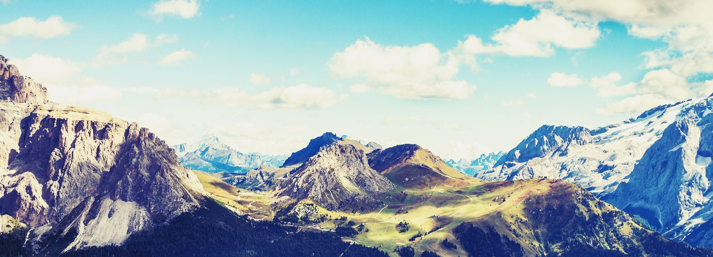 Mountainous landscape with rocky peaks, green valleys, and a bright blue sky with scattered clouds.