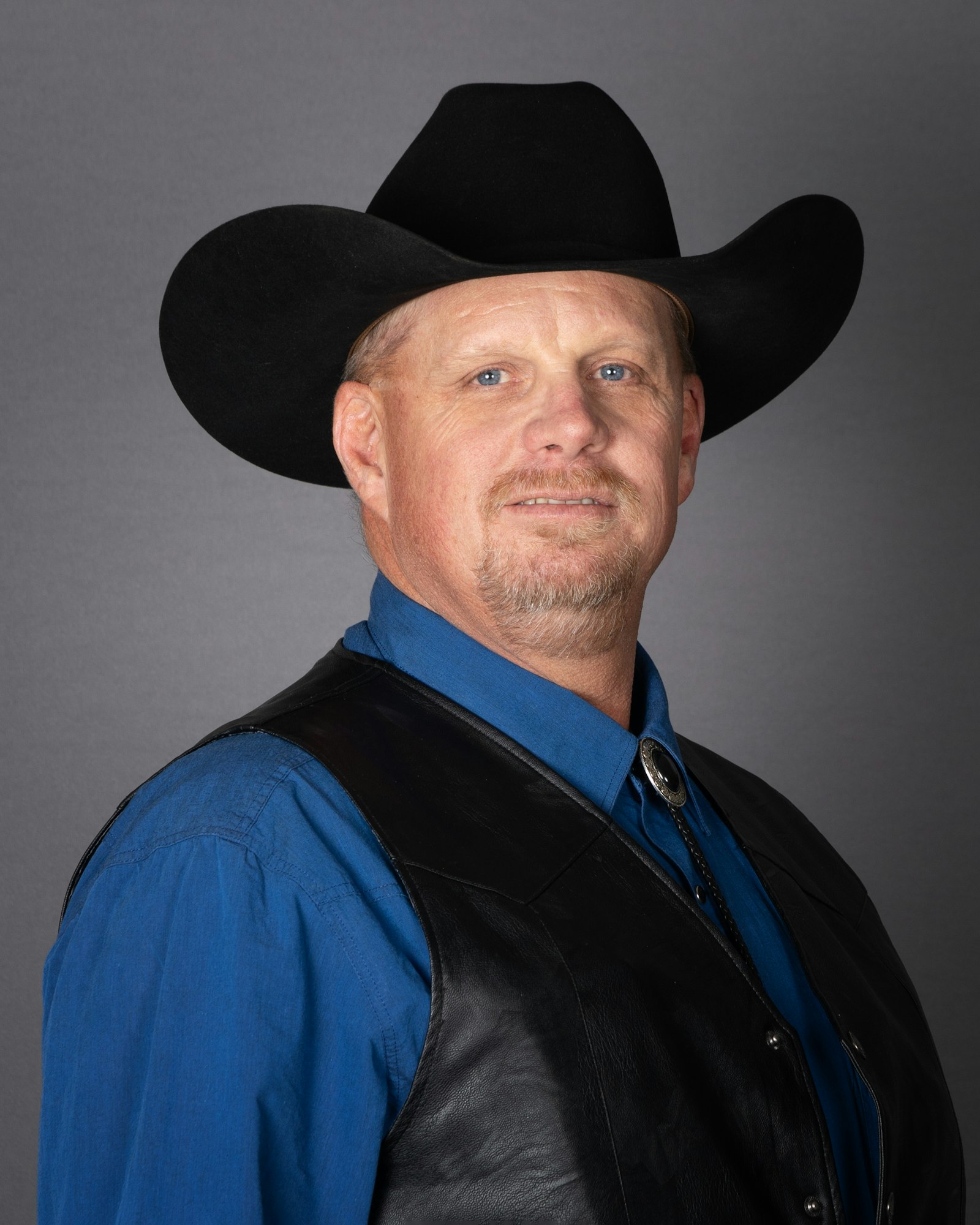 A man in a black cowboy hat and leather vest, with blue shirt, posed against a gray background.