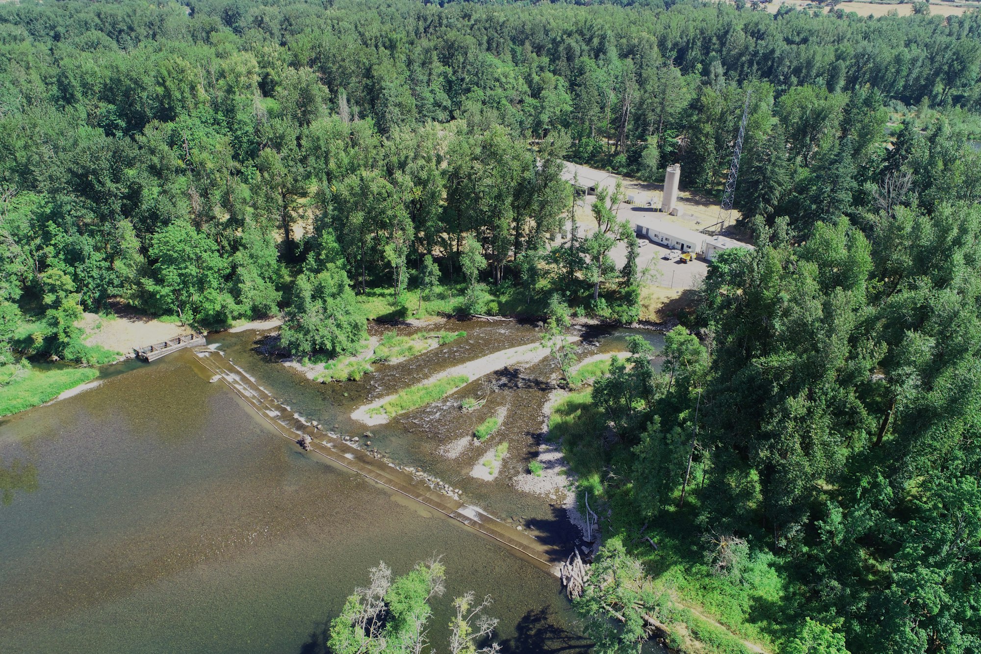 Aerial view of a river with a spillway, surrounded by dense forest and a building complex with a tall chimney.