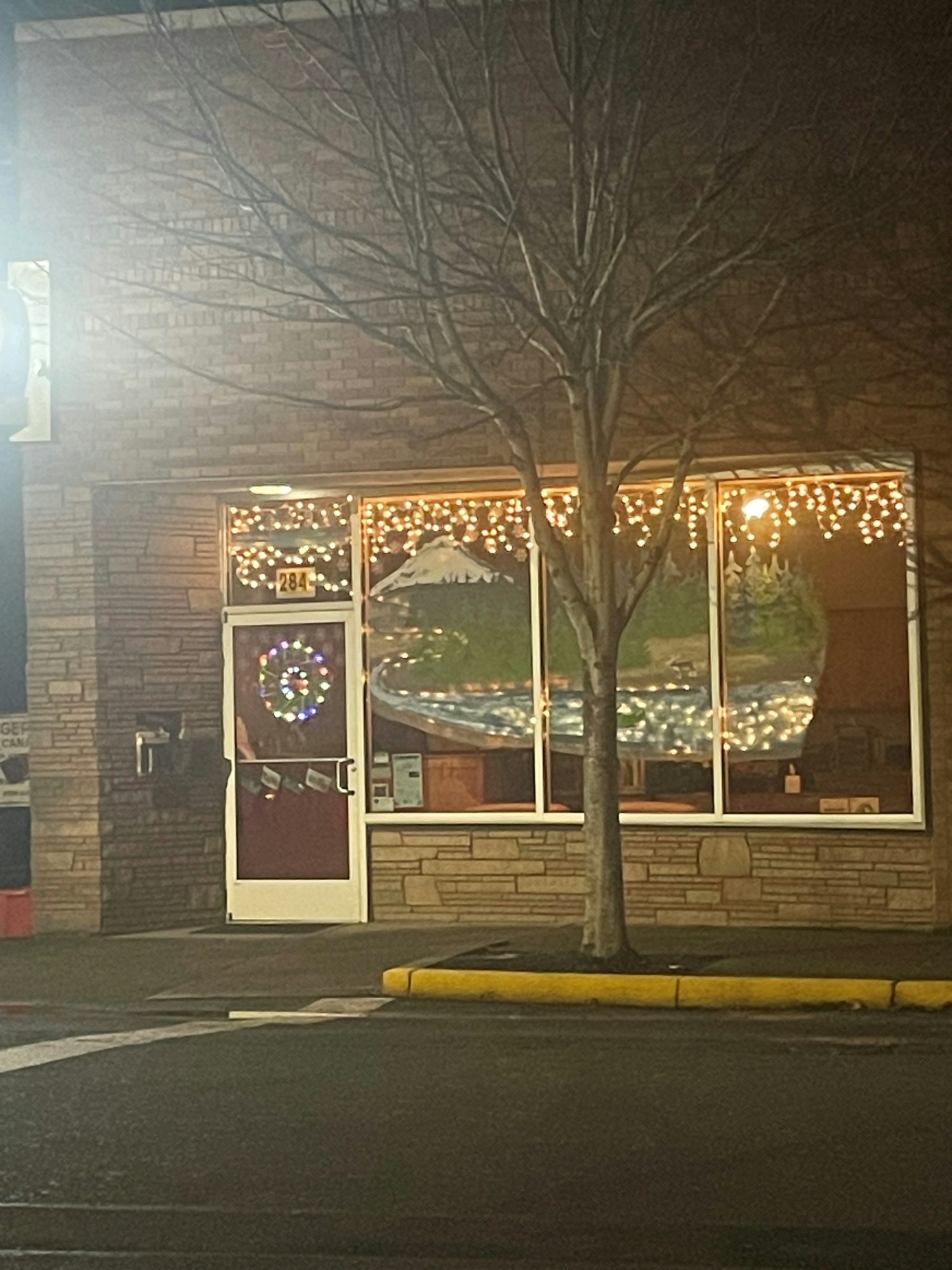 A brick building with a tree, window lights, and a door with a colorful wreath decoration.