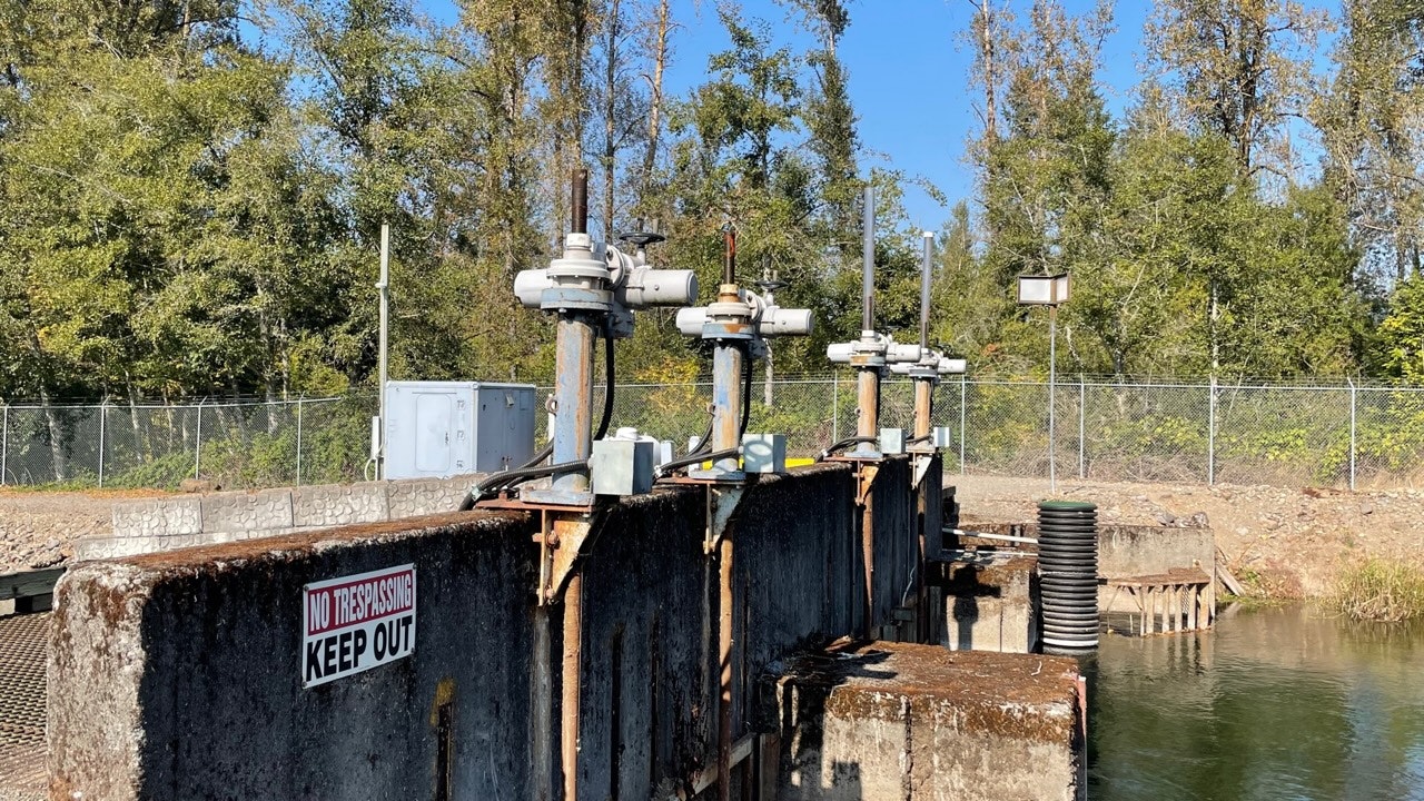 Industrial site with pipes and valves near water, "No Trespassing" sign visible, surrounded by fence and trees.