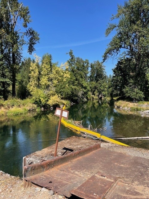 A serene river surrounded by lush trees with a rusty metal platform and sign in the foreground under a clear blue sky.