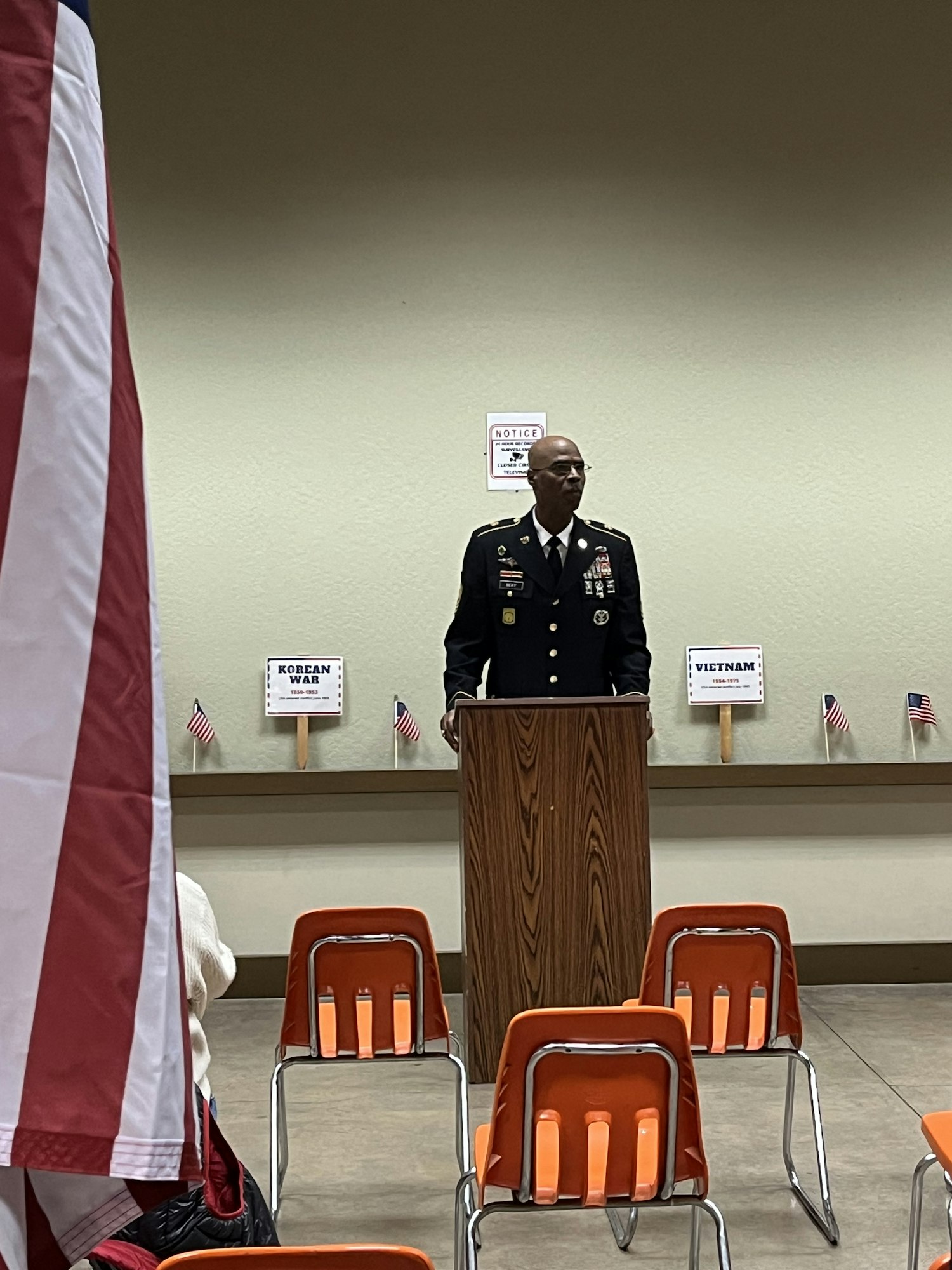 A person in uniform stands at a podium with small American flags and signs on the wall, orange chairs, and a large flag in the foreground.
