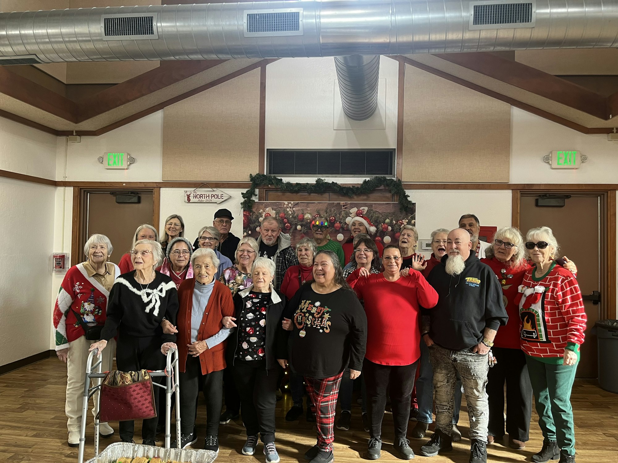A group of people in festive attire posing indoors, with holiday decorations and a "North Pole" sign in the background.