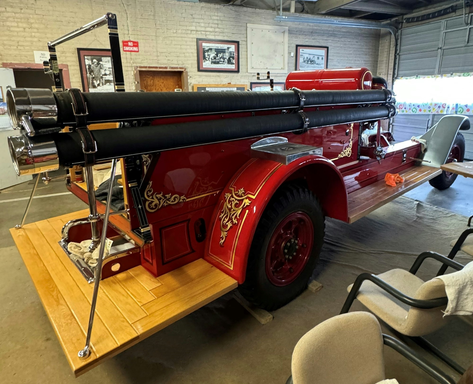A vintage red fire truck with decorative gold details, parked in a garage with no smoking sign and chairs nearby.
