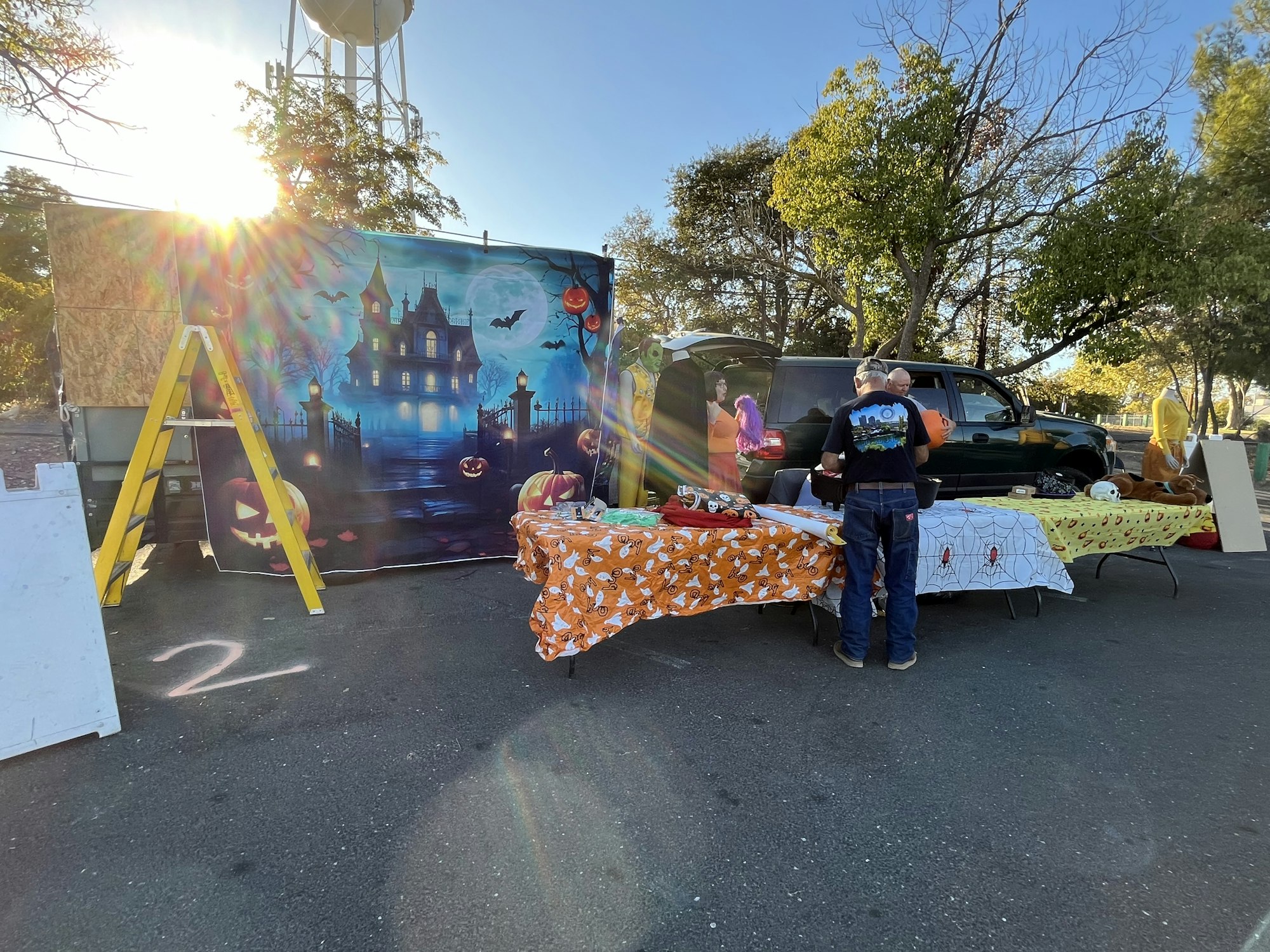 Outdoor Halloween setup with spooky backdrop, decorated tables with themed cloths, and people preparing items near a car.