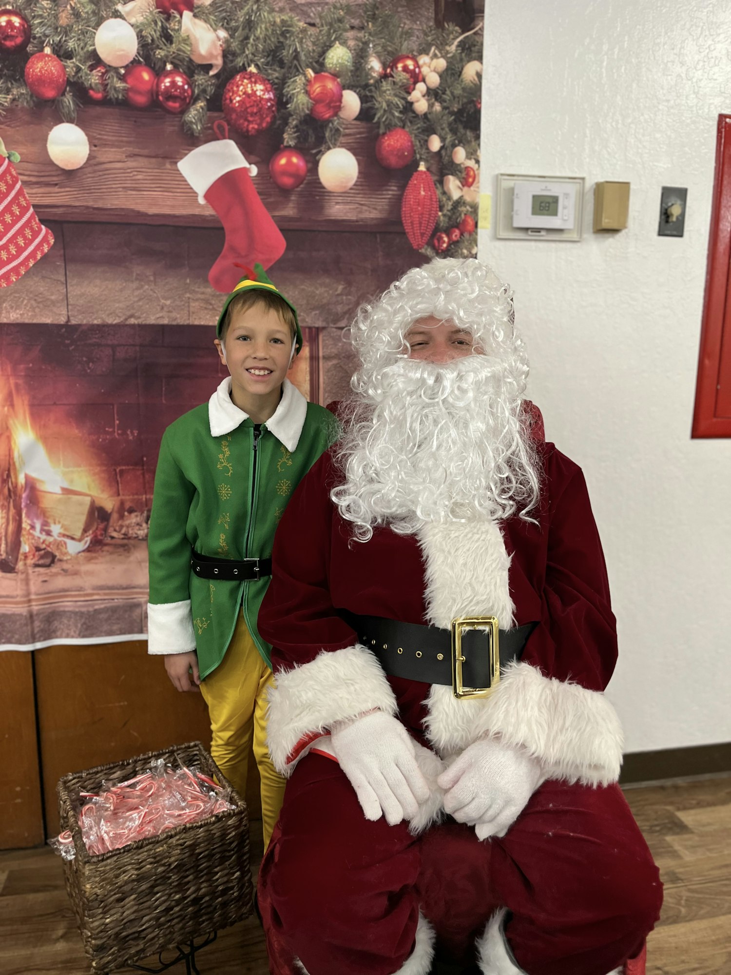 A person dressed as Santa Claus sitting next to a child in an elf costume, with a festive background and a basket of candy canes.