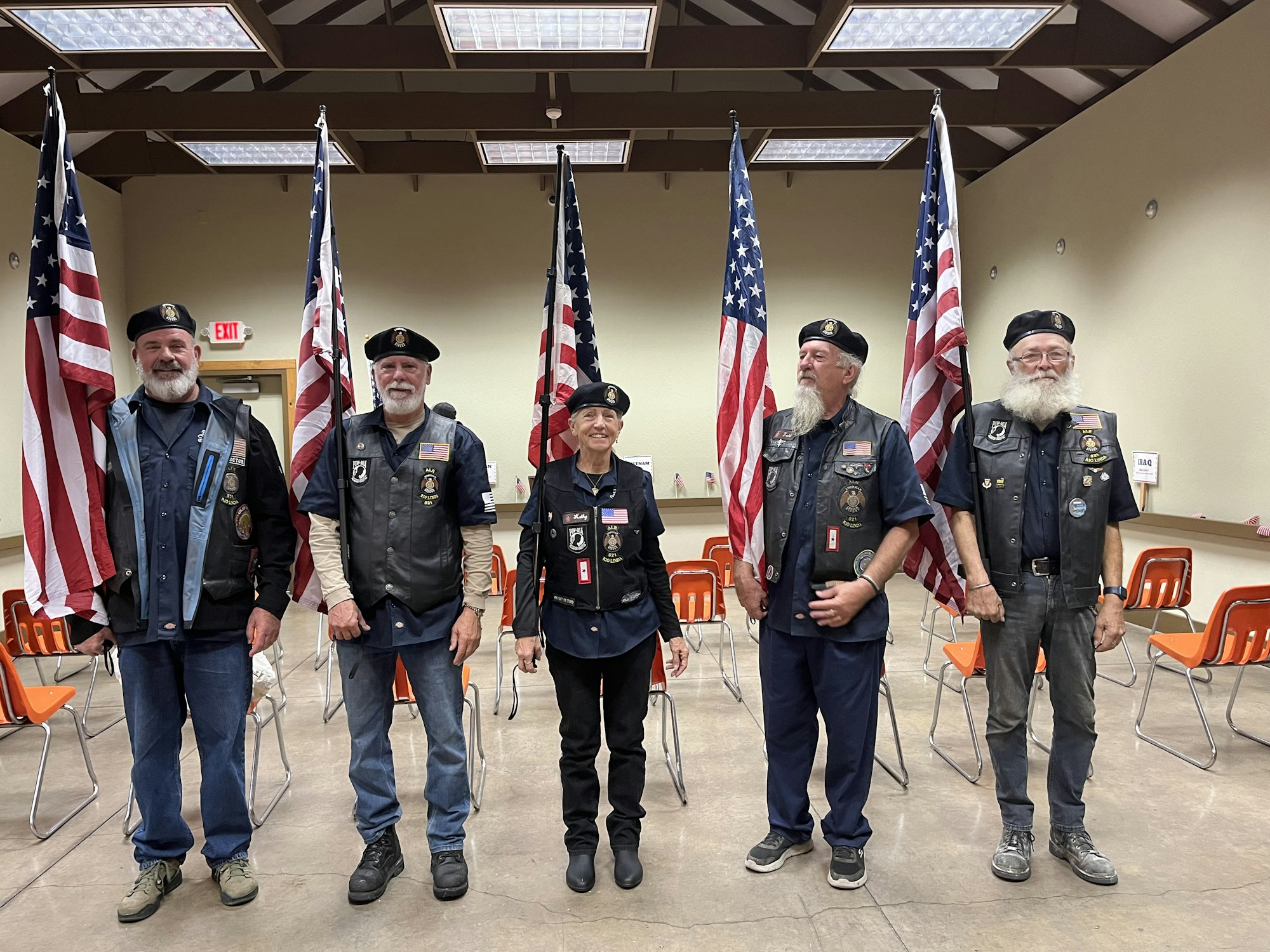 Five people in uniforms with patches, standing in front of U.S. flags in a room with orange chairs.