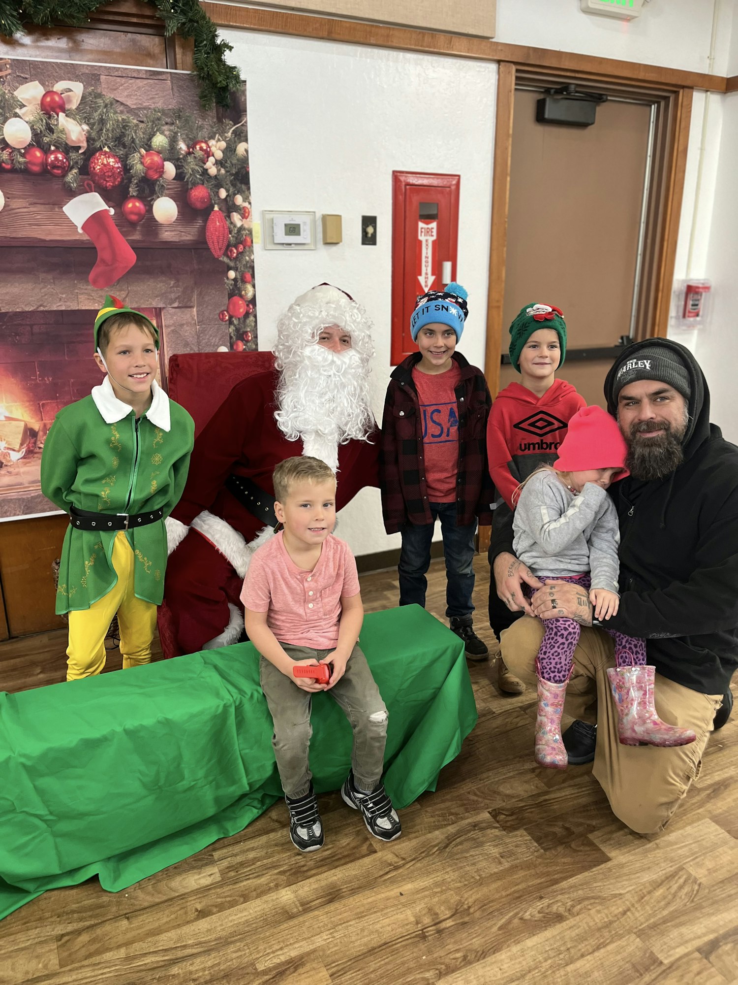 People posing with Santa, a kid dressed as an elf, holiday decorations, and a festive backdrop.